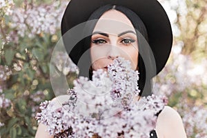 Pretty young woman with beautiful eyes in elegant stylish hat holds near face bouquet fresh lilac flowers on background flowering