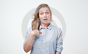 Pretty young woman in blue shirt pointing at herself in excitement being happy with success