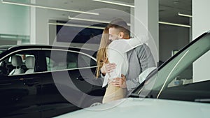 Pretty young woman is closing her husband`s eyes and leading him to new car in automobile showroom, excited man is