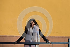 pretty young woman with curly brunette hair against a yellow background is dressed in winter clothes and wearing a scarf to