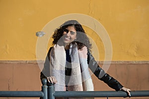 pretty young woman with curly brunette hair against a yellow background is dressed in winter clothes and wearing a scarf to