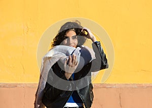 pretty young woman with curly brunette hair against a yellow background is dressed in winter clothes and wearing a scarf to