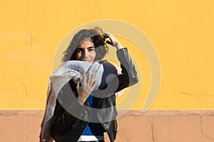 pretty young woman with curly brunette hair against a yellow background is dressed in winter clothes and wearing a scarf to