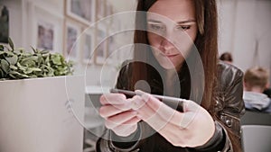 Pretty young woman with dark hair in a leather jacket using her smartphone, talking, smiling. Pictures on the wall.