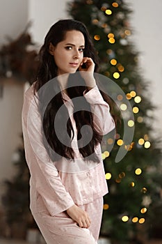 Pretty young woman with dark hair wearing modish sleepwear posing near decorated Christmas tree