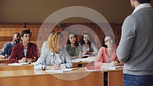 Pretty young woman diligent student is talking to high school teacher sitting at desk while other students are smiling