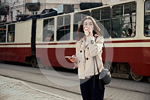 Pretty young woman drinking takeout coffee cup, using smart phone while waiting train as arrival.