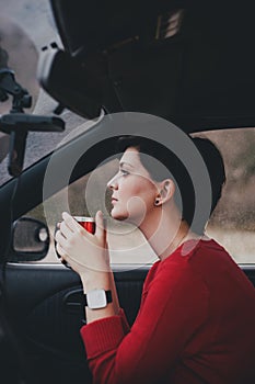 Pretty young woman drinking tea in a car with rain outside