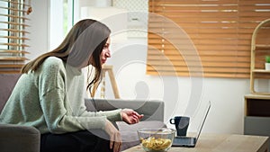 Pretty young woman eating cookies while talking on video call with girl friend via laptop at home