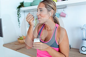 Pretty young woman eating yogurt while listening to music with wireless earphones in the kitchen at home