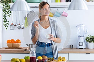 Pretty young woman eating yogurt while standing in the kitchen at home