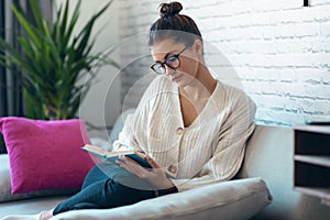 Pretty young woman with eyeglasses reading a book while sitting on sofa at home