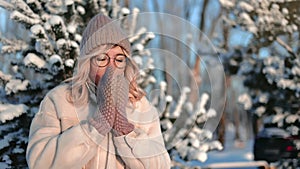 Pretty young woman feeling cold in forest in winter day, warming hands in knitted mittens, portrait