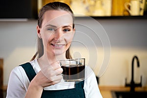 Pretty young woman in a green kitchen apron with a cup of coffee in her hands is sitting in the kitchen and relaxing.