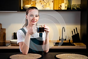 Pretty young woman in a green kitchen apron with a cup of coffee in her hands is sitting in the kitchen and relaxing.