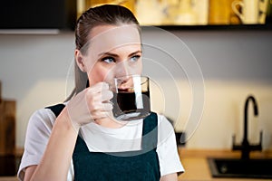 Pretty young woman in a green kitchen apron with a cup of coffee in her hands is sitting in the kitchen and relaxing.