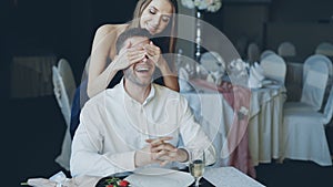 Pretty young woman is greeting her boyfriend in restaurant closing his eyes with her hands