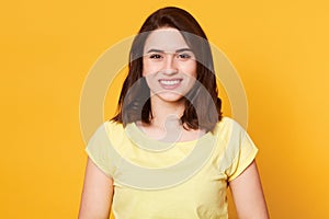 Pretty young woman with happy facial expression, stands smiling against yellow studio wall, female has brown hair, dressed