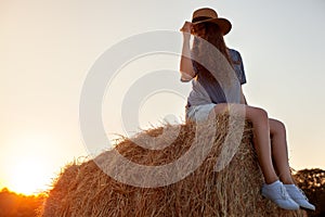 Pretty young woman in hat sitting on a hay bale