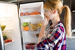 Pretty young woman hesitant to eat in front of the fridge in the kitchen.