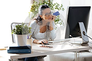 Pretty young woman holding blue credit card for shopping online with computer at home