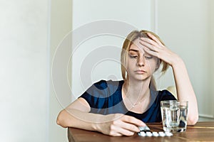 Pretty young woman holding his head and feeling sick. The girl is sitting at the table and looking at pain medication