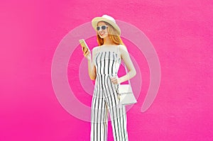 pretty young woman holding smartphone in summer round hat, white striped jumsuit on colorful pink wall