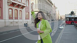 Pretty young woman holding takeaway coffee and calling a taxi on the street