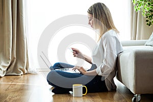 Pretty young woman holding white credit card for shopping online with computer while sitting on the floor at home