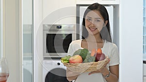 Pretty young woman holding wicker basket with fresh organic vegetables and fruits standing in home kitchen