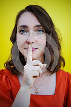 Pretty young woman keeps finger on lips, making hush gesture on yellow background. Shh, silence concept.