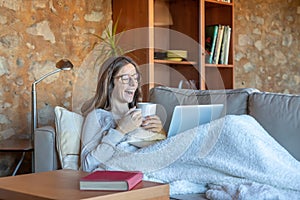 Pretty young woman laughing sitting on the sofa at home looking at the laptop screen and drinking a tea.