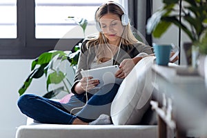 Pretty young woman listening to music with headphones and her digital tablet while sitting on sofa at home