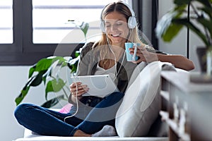 Pretty young woman listening to music with her digital tablet while drinking cup of coffee on sofa at home