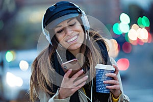 Pretty young woman listening to music with mobile phone while drinking coffee in the street at night