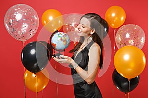 Pretty young woman in little black dress celebrating, looking on world globe in hands on red background air balloons