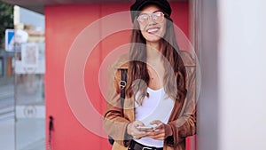 Pretty young woman looking at camera while using her mobile phone in the street.