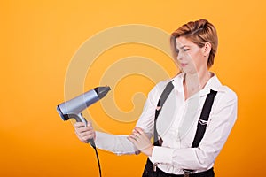 Pretty young woman looking confused at her hair drayer over yellow background.