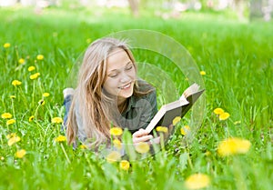 Pretty young woman lying on grass with dandelions reading a book