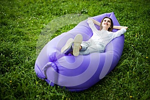 Pretty young woman lying on inflatable sofa lamzac while resting on grass in park on the sun under sky
