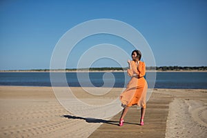 Pretty young woman in an orange dress, posing on a wooden catwalk on the beach, with the wind ruffling her hair and dress. Concept