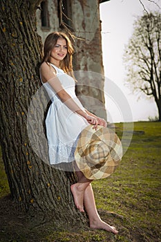 Pretty young woman posing in front of the farm. Very attractive blonde girl with white short dress holding a hat. Romantic girl
