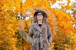 Pretty young woman with a positive smile in a stylish coat in a fashionable hat in trendy glasses stands in the park and enjoys