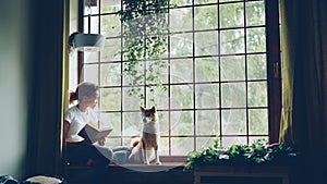 Pretty young woman is reading book sitting on windowsill and looking outside together with cute pedigree dog. Beautiful