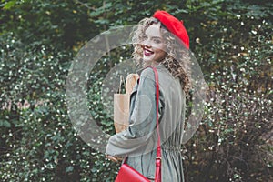 Pretty young woman in red beret holding craft paper bag with fresh french baguette against green leaves