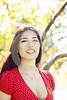 Pretty young woman in red dress smiling cheerful in green park at tree on summer sunny day, lifestyle people concept