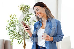 Pretty young woman sending messages with mobile phone while eating yogurt at home