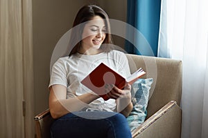 Pretty young woman sits at comfortable armchair, reads interesting book, dressed in white casual t shirt and jeans, poses near