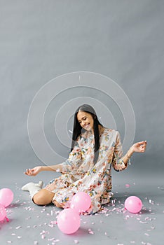 A pretty young woman sits near pink balloons and throws up confetti and smiles and on a gray background