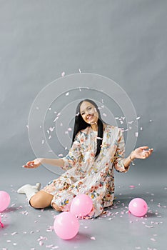 A pretty young woman sits near pink balloons and throws up confetti and smiles and on a gray background